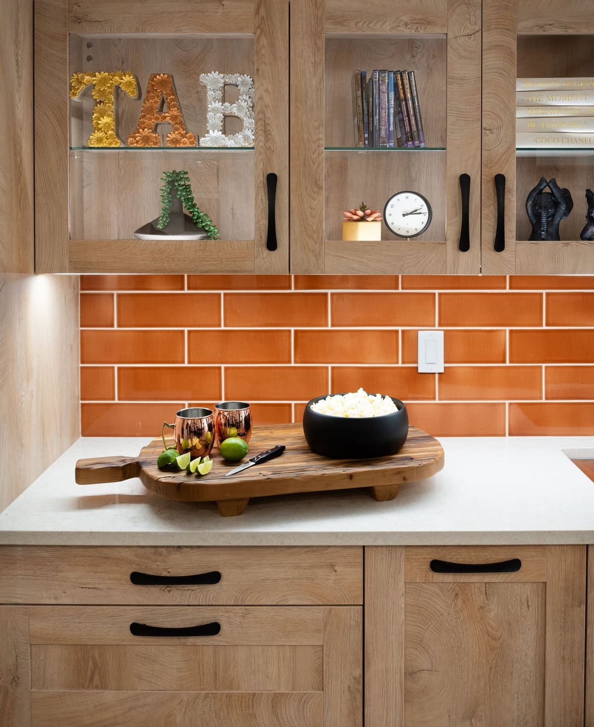 Kitchenette vignette with copper mugs and popcorn on a wood board below glass-front light oak cabinets