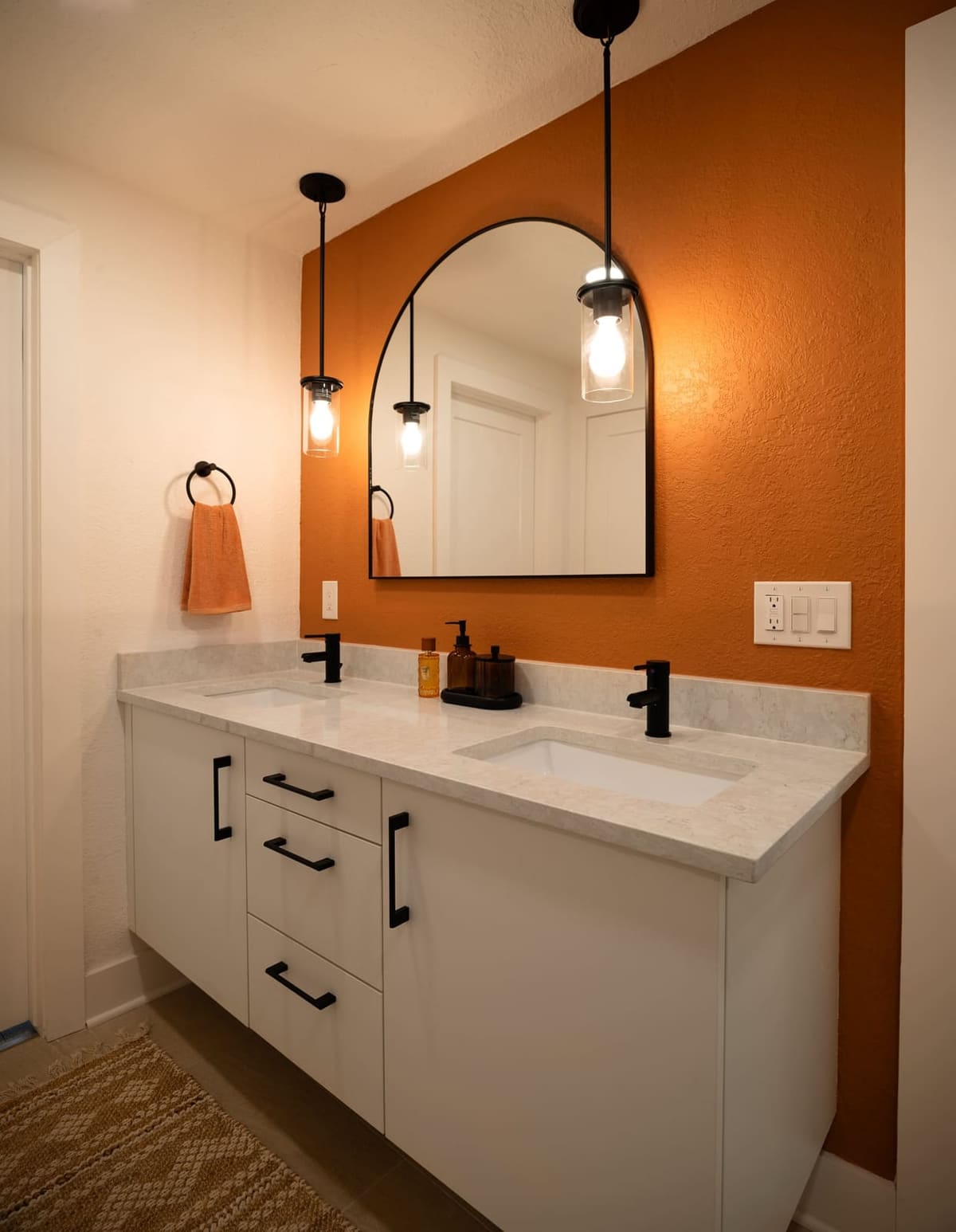Bath with an orange accent wall, white double vanity, arched black mirror and glass pendants