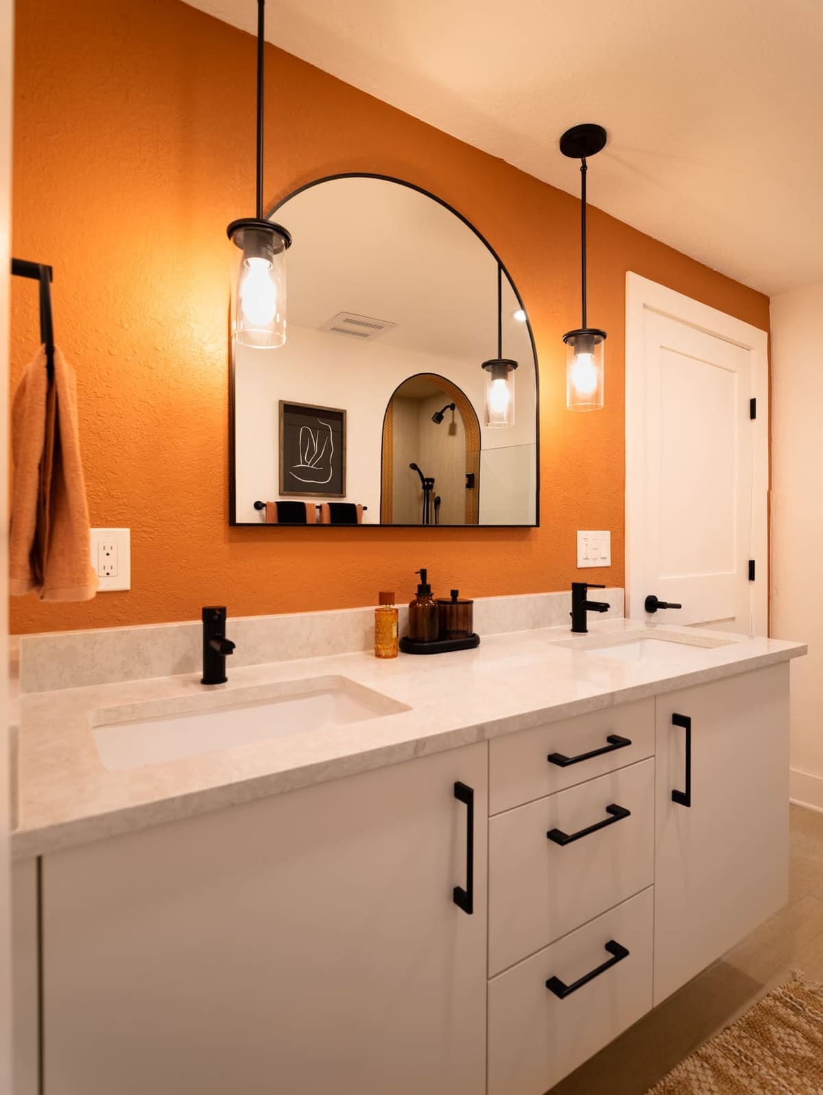 White double vanity with black pulls under an arched black mirror on an orange wall