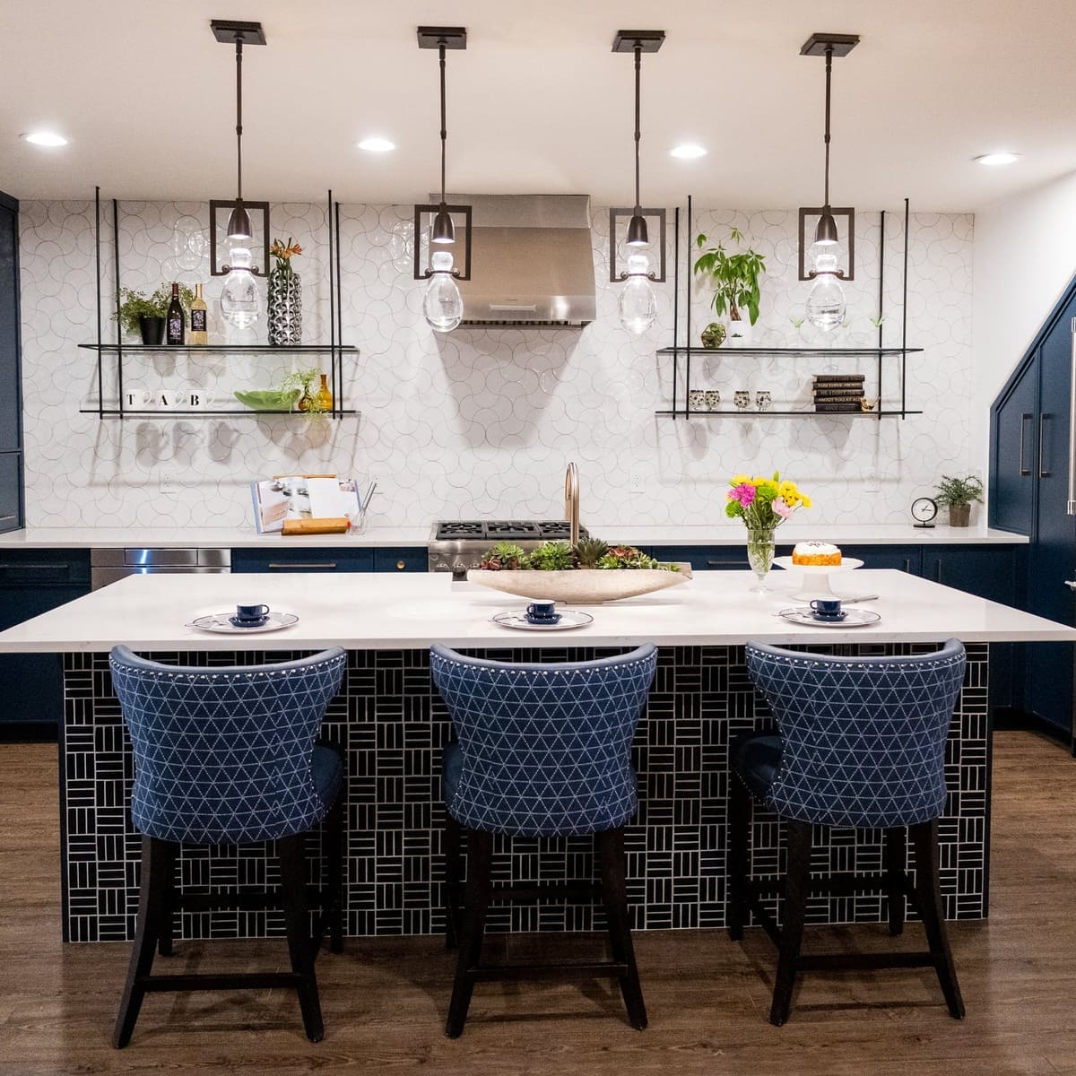 Basement kitchen with a navy island, blue nailhead stools and glass pendants under hanging metal shelves