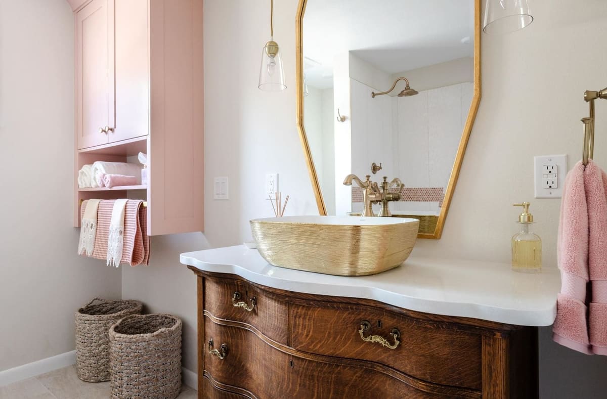 Antique wood dresser vanity with a gold vessel sink, pink cabinet and gold-framed mirror