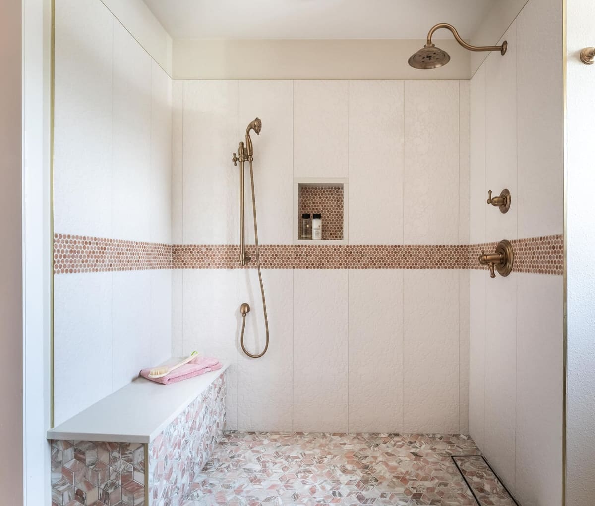 Walk-in shower with white tile, a pink pebble band, tiled bench and brushed gold fixtures