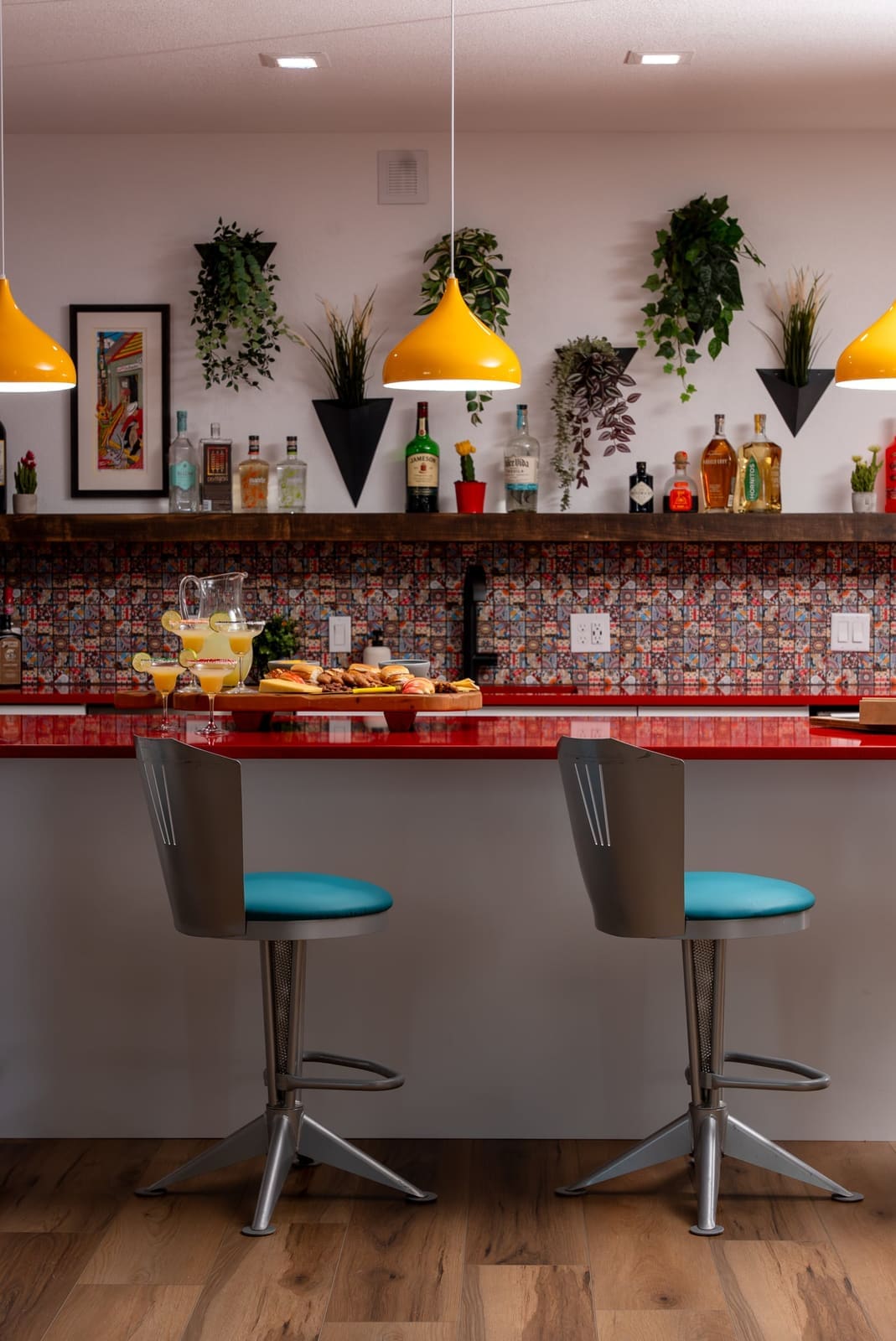 Home bar with two swivel stools with teal seats, yellow dome pendants, red quartz and patterned tile