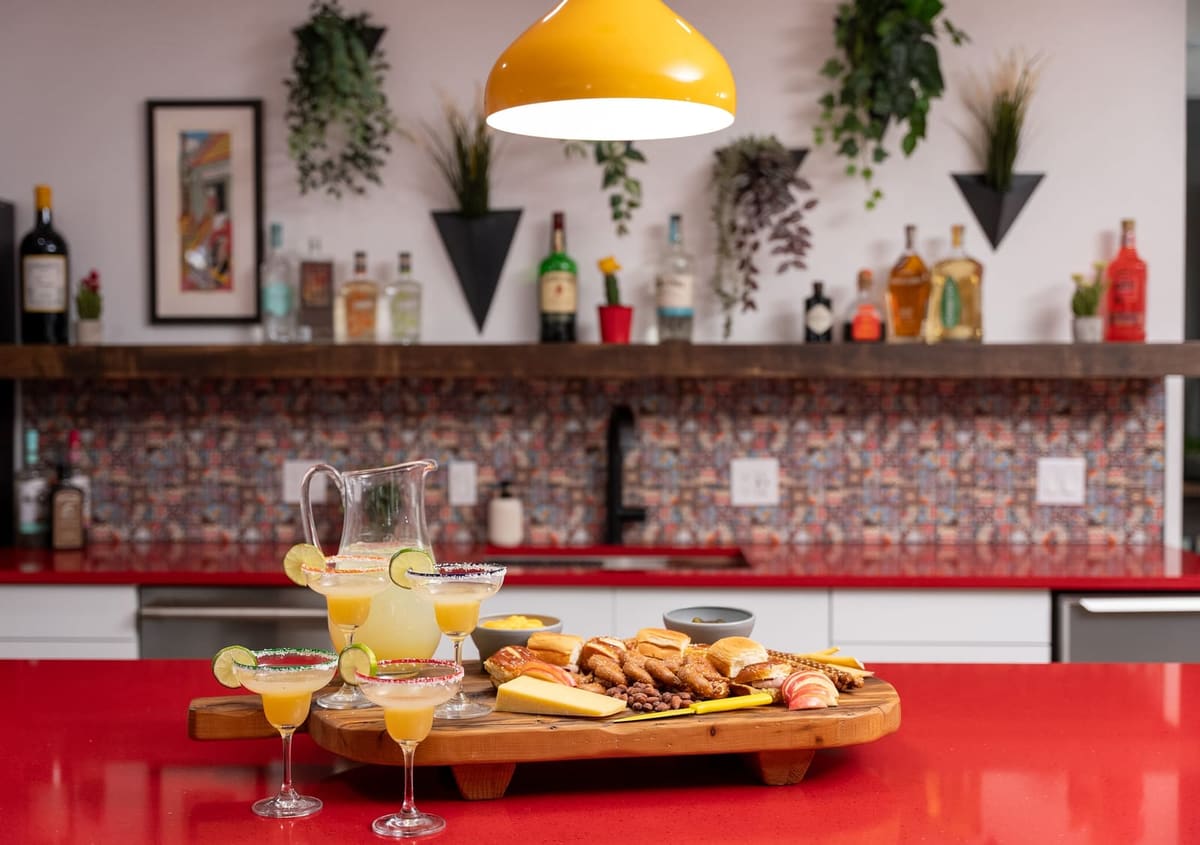 Home bar with a red quartz counter, yellow pendant, margaritas and a wood shelf above patterned tile