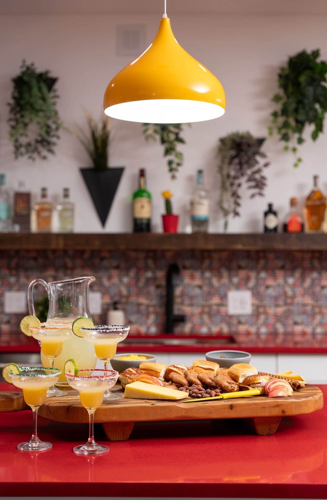 Margaritas and a charcuterie board on a red quartz bar under a yellow pendant with wall planters beyond