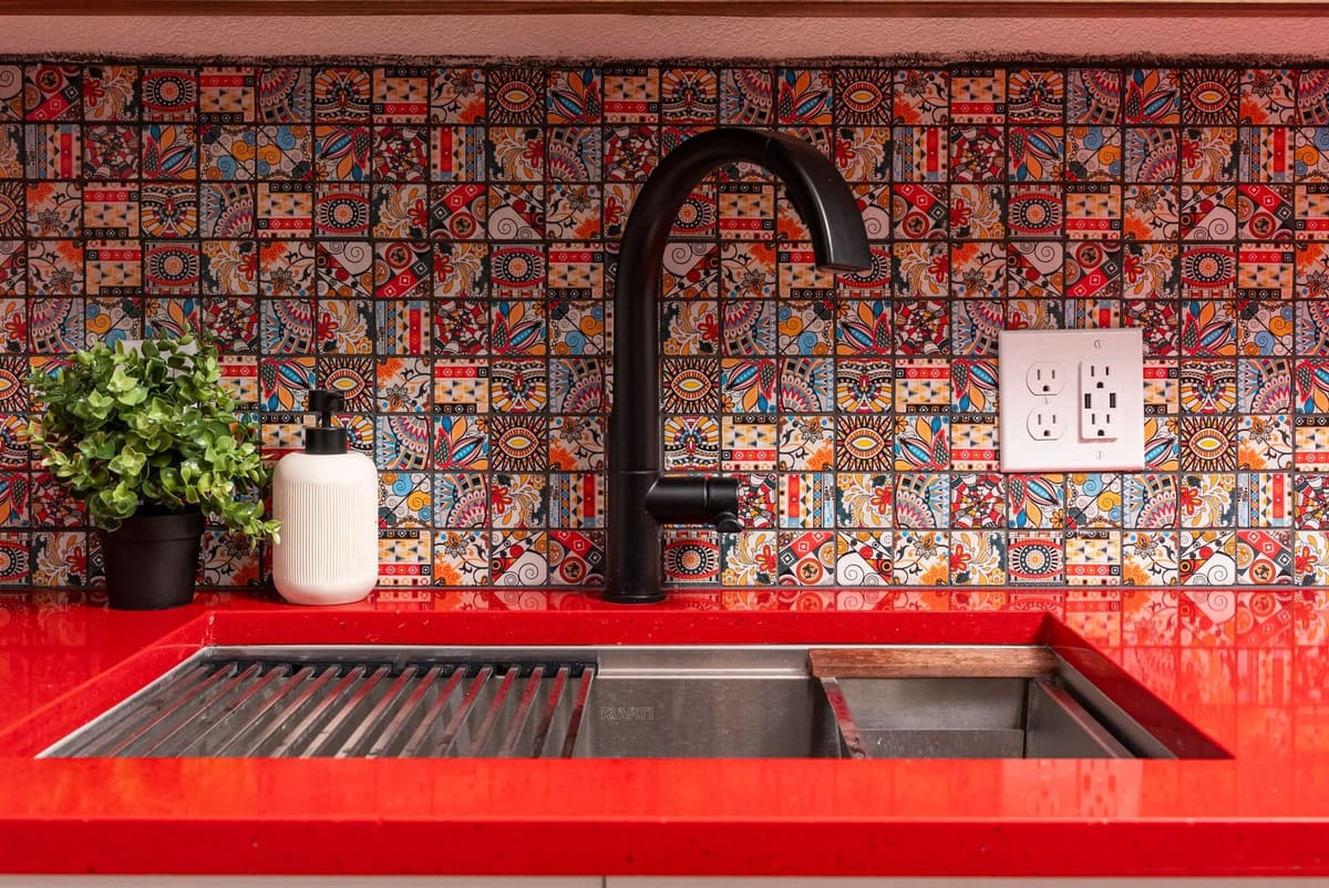 Matte black faucet over a workstation sink in red quartz with a multicolor patterned mosaic backsplash