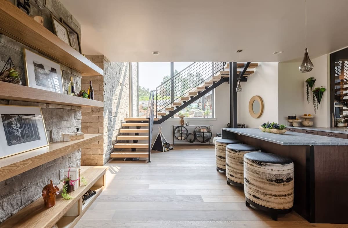 Open oak stair with black steel railing beside a wet bar island with gray stone top and upholstered drum stools