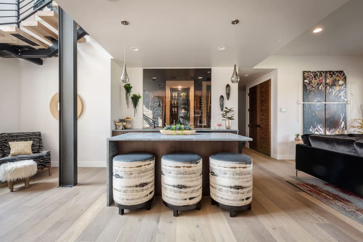 Wet bar island with three drum stools, glass teardrop pendants, dark wood cabinetry and a lit wine display behind