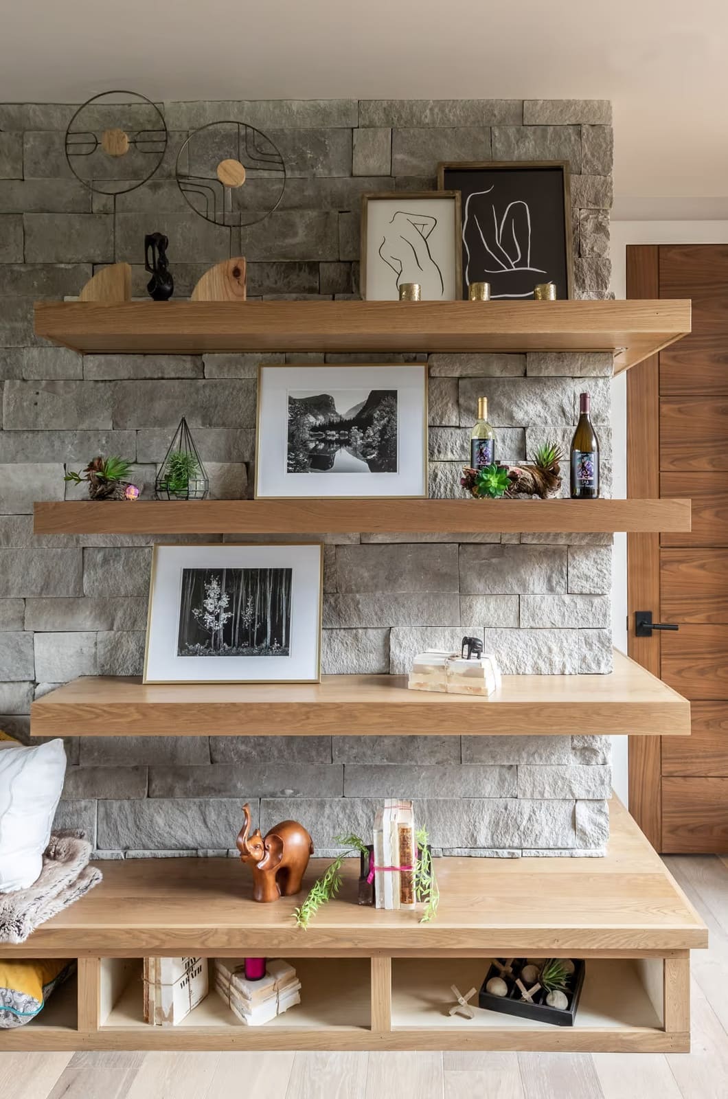 White oak floating shelves on gray stacked stone with framed black-and-white photos, line drawings and wine bottles