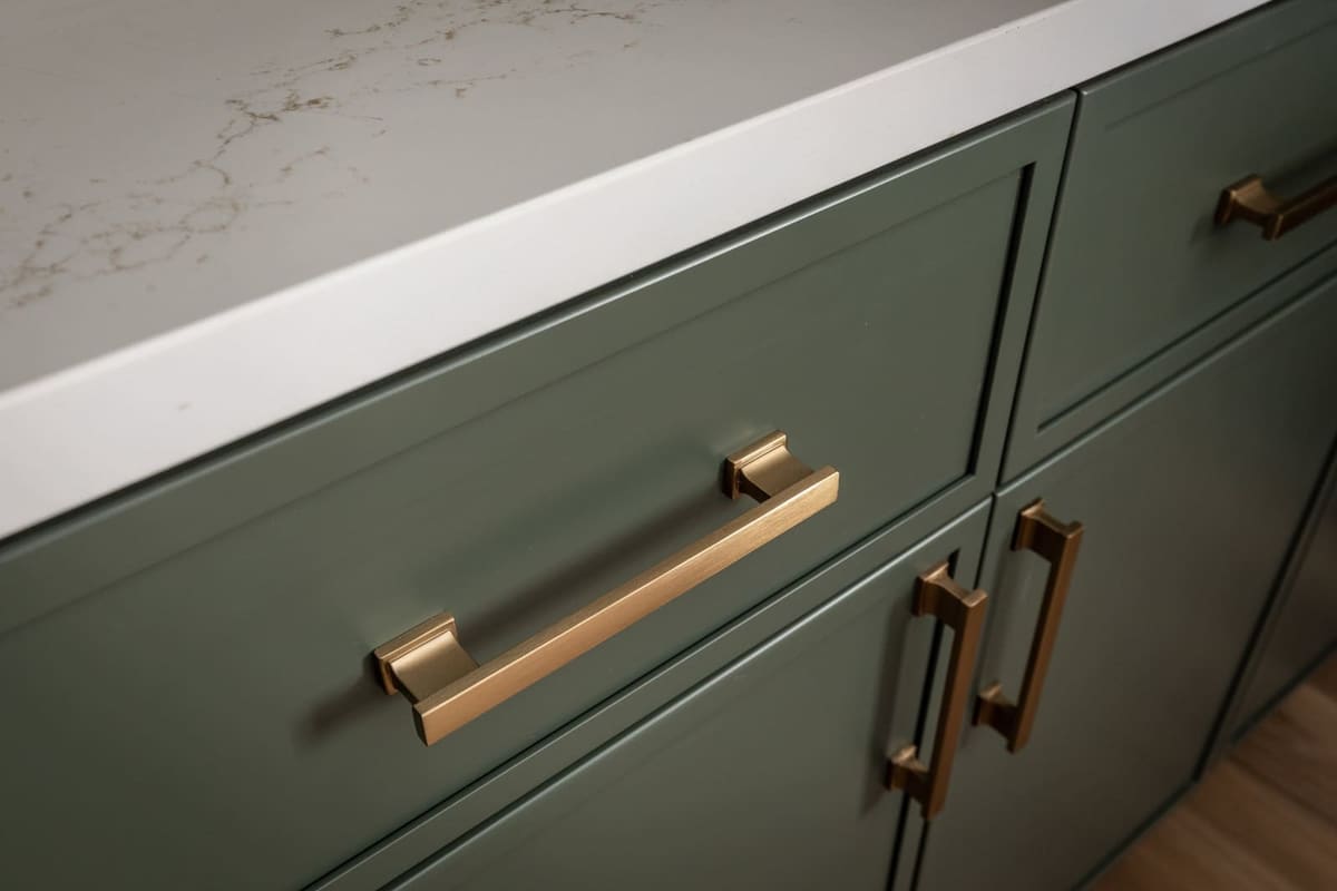 Brushed brass pulls on sage green shaker drawers under a white quartz edge