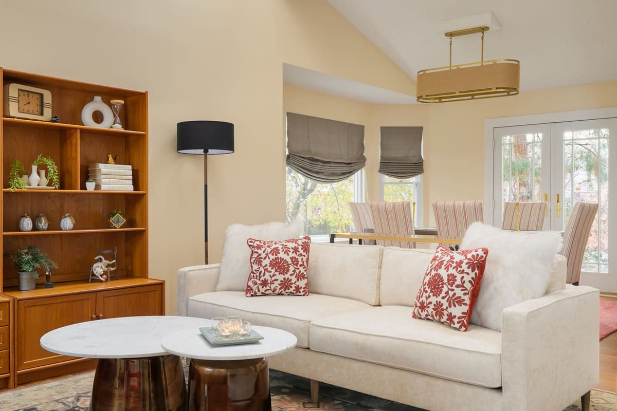 Living room with a cream sofa, red floral pillows, marble nesting tables and a teak bookcase, dining beyond
