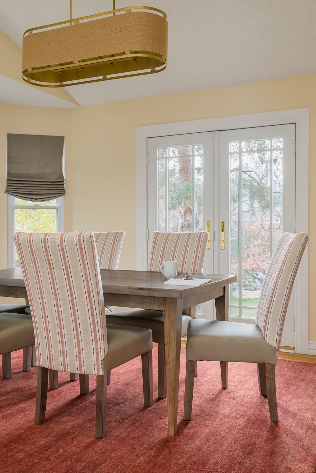 Dining room with striped high-back chairs, a gray wood table, red rug and an oval rope and brass chandelier