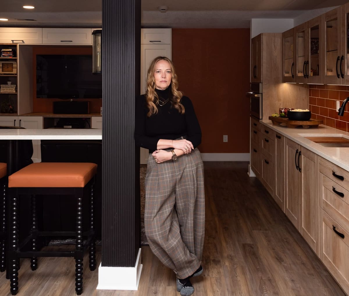Trish Bonney leaning on a black fluted column in a finished basement bar with oak cabinets and orange tile